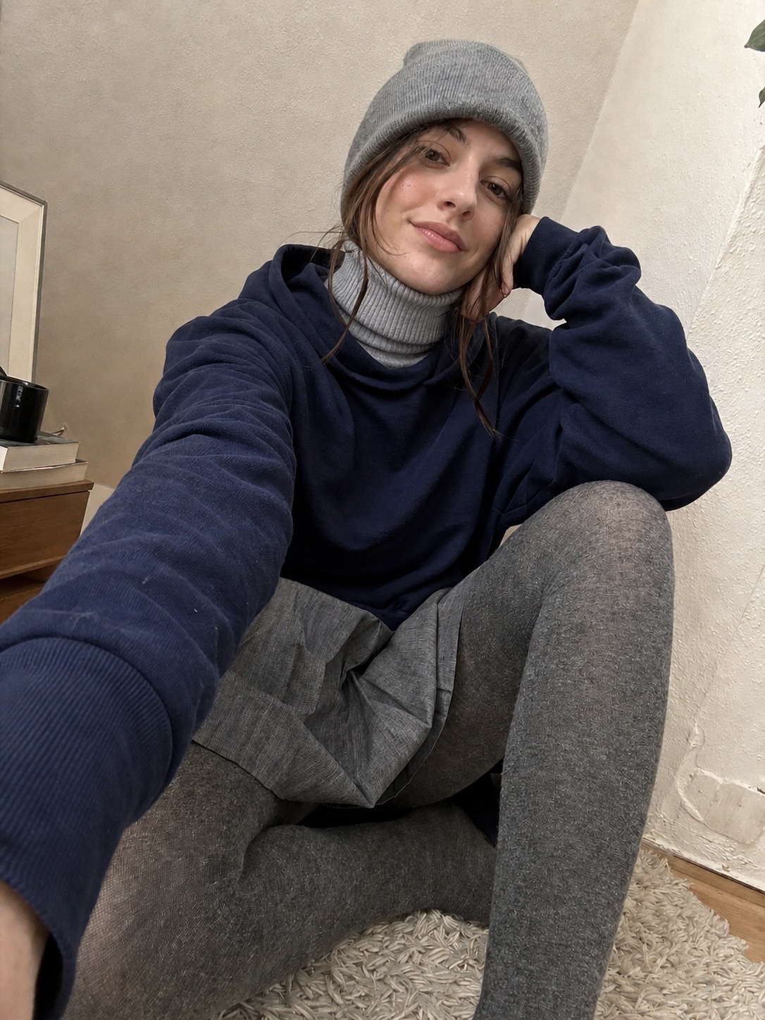 Frau sitzt entspannt auf einem Teppich und trägt graue Strickstrumpfhose, Hoodie und Mütze in ruhiger Wohnatmosphäre